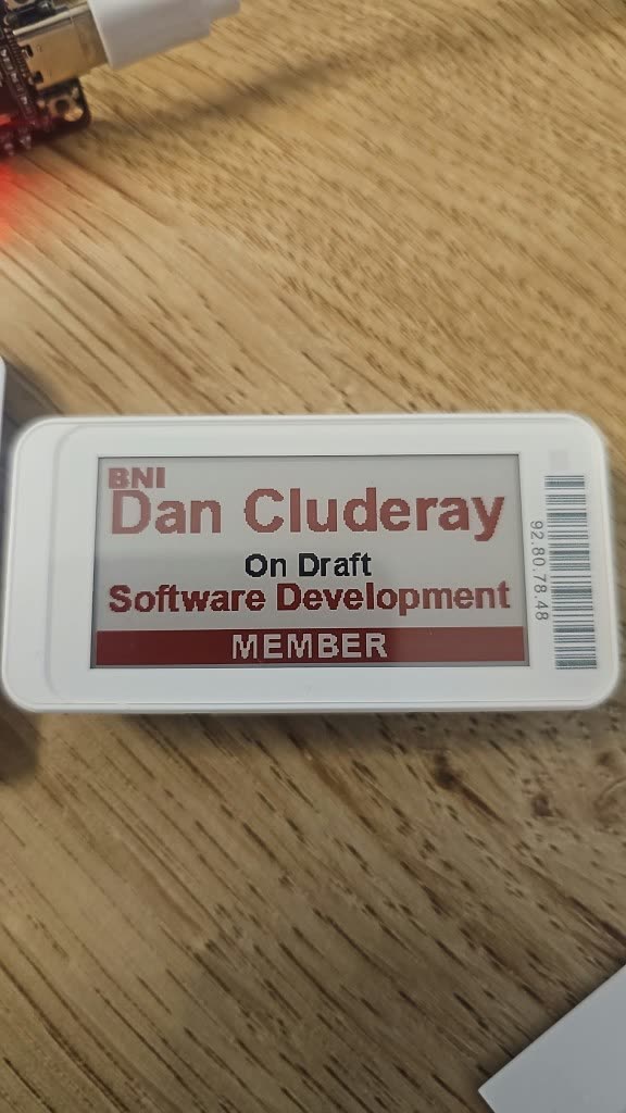 Physical e-ink badge on a wooden desk with a USB-C cable attached